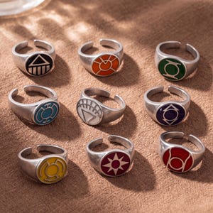 May include: A collection of nine silver-toned metal rings, each featuring a circular face with a unique colored enamel inlay and distinct geometric symbol, including black, orange, green, blue, white, purple, yellow, maroon, and red designs.