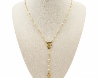May include: A gold-toned rosary necklace featuring small white beads spaced along a delicate chain. The necklace includes a central oval-shaped medal depicting a figure and terminates in a simple gold-toned cross pendant at the bottom.