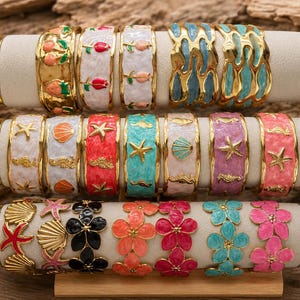 Peut inclure: Une collection de dix-huit bracelets manchette en métal présentés sur trois supports cylindriques. Les bracelets présentent divers motifs en émail, notamment des fleurs, des coquillages, des étoiles de mer, des hippocampes et des motifs abstraits aux couleurs vives comme le rose, le bleu, l'orange et le blanc.