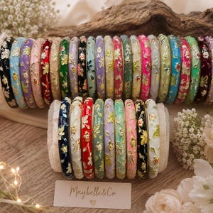 Peut inclure: Une collection de bracelets joncs fins en métal de différentes couleurs, dont noir, bleu, rose, rouge, vert et blanc, chacun orné d'un motif floral doré délicat. Un petit panneau rectangulaire devant affiche le texte Maybelle & Co avec un cœur.