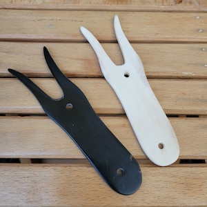 May include: Two yarn needle tools, one black and one white, with a wooden handle and two prongs. The tools are on a wooden surface.