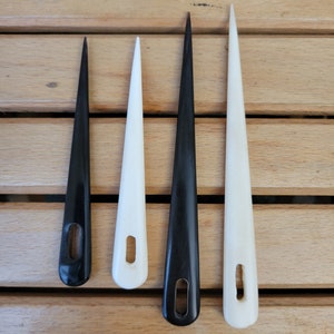 May include: Four needle tools, two white and two black, with a small hole near the top of each needle. The needles are arranged in a row on a wooden surface.