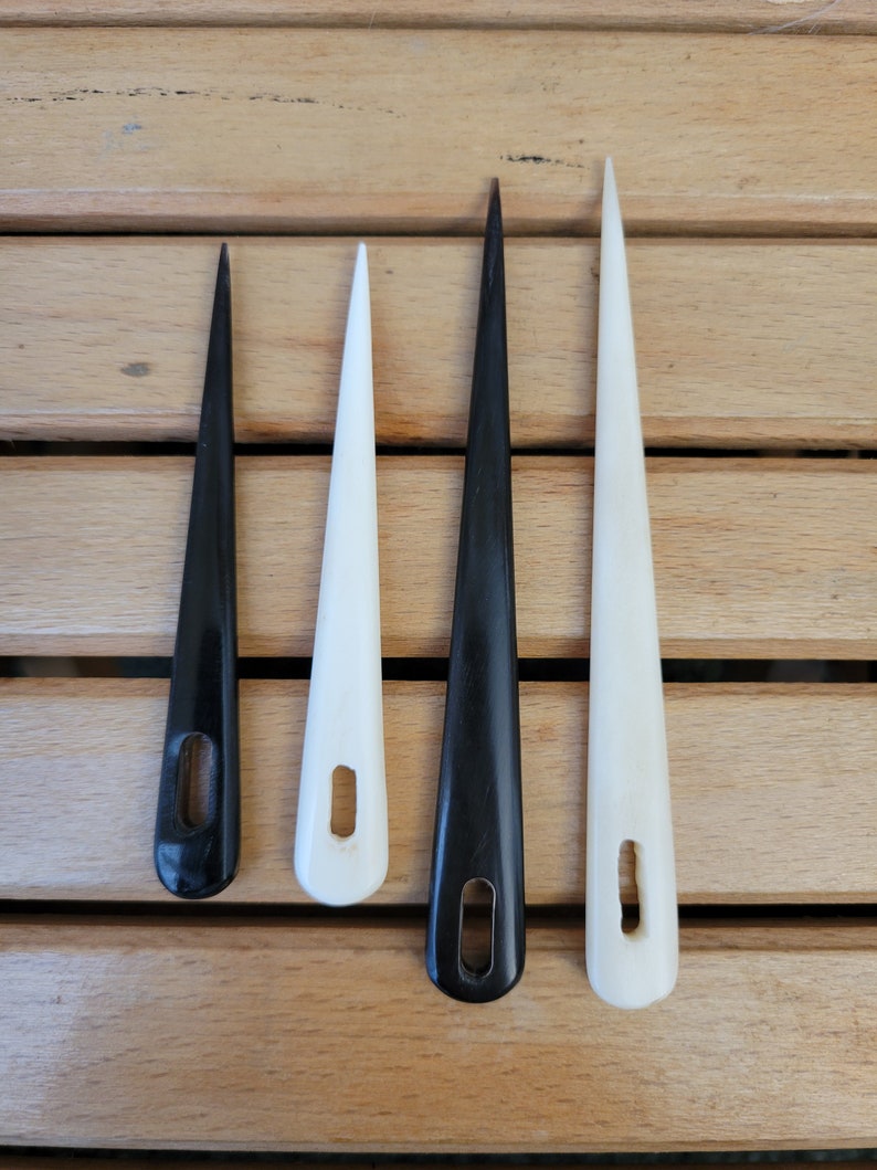 May include: Four needle tools, two white and two black, with a small hole near the top of each needle. The needles are arranged in a row on a wooden surface.