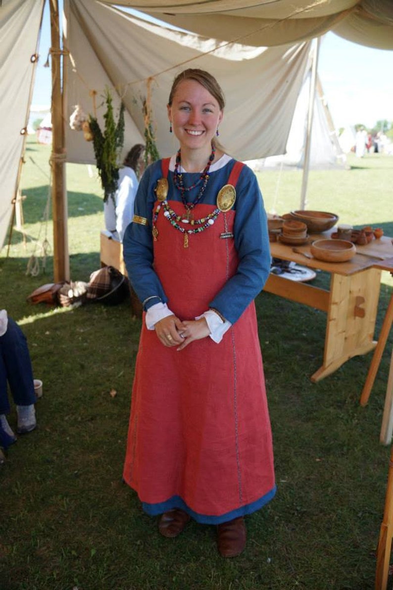 Op de afbeelding: Een vrouw draagt een blauwe tuniek over een rode jurk met een kralenketting en decoratieve broche. Ze staat voor een witte tent.