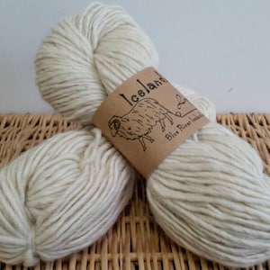 Puede incluir: Un ovillo de lana islandesa blanca, con una etiqueta que dice "Icelandic Blue River Yarns". La lana está sobre una superficie de mimbre.