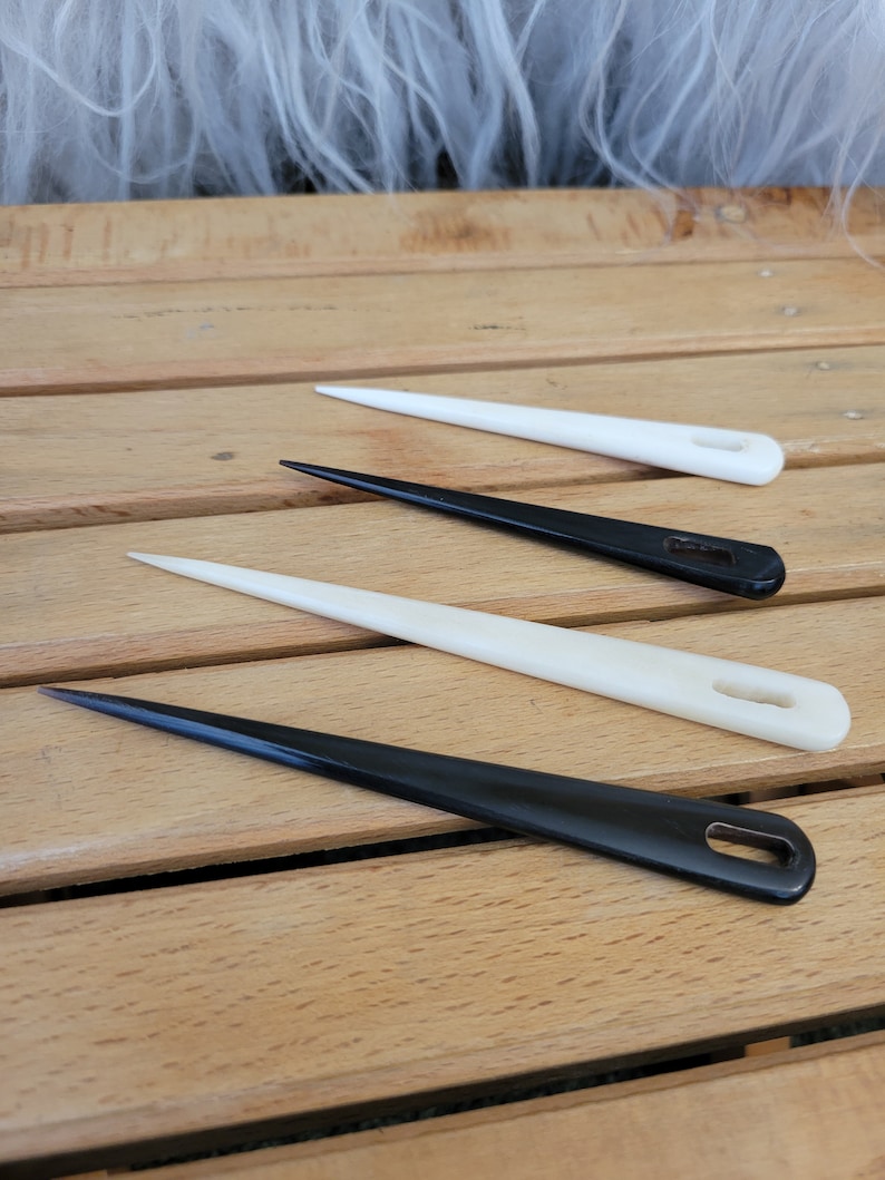 May include: Three leather needles, two white and one black, with a pointed tip and a small hole at the end. The needles are laid out on a wooden surface.