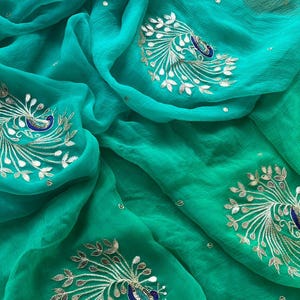 May include: A vibrant teal fabric with embroidered peacock motifs. The embroidery uses silver-coloured thread to depict the peacock's feathers and body, with blue accents. The fabric is draped, highlighting its texture and the detailed craftsmanship of the design.