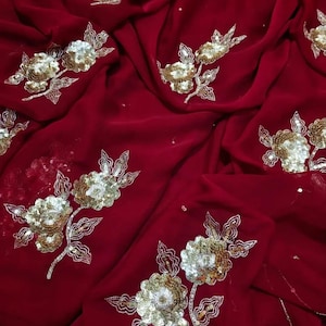 May include: A rich crimson fabric adorned with embroidered floral designs. The flowers feature gold sequins and silver detailing. The fabric is draped, highlighting the texture and pattern of the material.