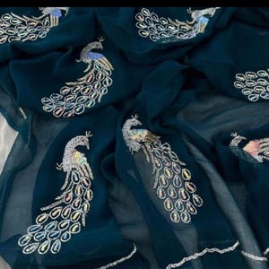 May include: Dark teal fabric with embroidered peacock designs. The peacock feathers are embellished with iridescent beads. The fabric is draped, showing the intricate details of the embroidery. The overall design is elegant and decorative.