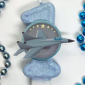 Puede incluir: Una vela de cumpleaños con forma de número uno en color azul claro brillante, con un emblema circular que muestra un avión de combate gris y cinco estrellas amarillas. La vela está rodeada por un collar de cuentas azules y blancas.