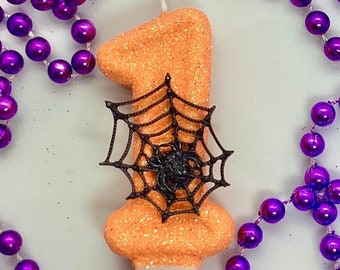 May include: An orange glitter candle in the shape of the number one, decorated with a black spider web and spider. The candle is surrounded by purple beaded necklaces, perfect for a Halloween or birthday celebration.