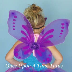 May include: A young girl wearing a blue, purple and pink tutu with butterfly wings. The wings are purple with a light purple outline and have a flower in the centre. The tutu is made of tulle and has a layered design. The girl is wearing sandals and has her hair pulled back in a ponytail.