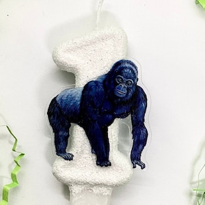 Può includere: Candela di compleanno bianca a forma di numero uno con finitura glitterata. Un adesivo di gorilla è attaccato alla candela.