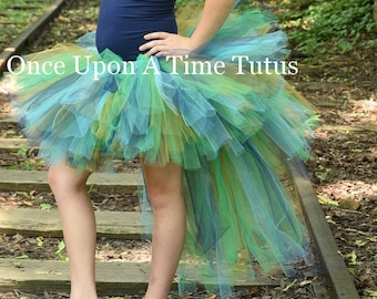 May include: A young person wearing a blue tank top and a multi-colored tulle tutu with a long train. The tutu has shades of green, blue, and gold. The person is standing on a set of railroad tracks in a wooded area. The text "Once Upon A Time Tutus" is visible on the tutu.