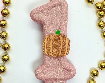 May include: A pink glitter candle in the shape of the number one, decorated with a glittery orange pumpkin. The candle is surrounded by strands of gold and magenta beads, perfect for a first birthday celebration.