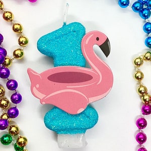 Puede incluir: Una vela de cumpleaños de color azul turquesa y rosa en forma de flamenco con el número uno en ella. La vela está rodeada de una guirnalda de cuentas multicolores.