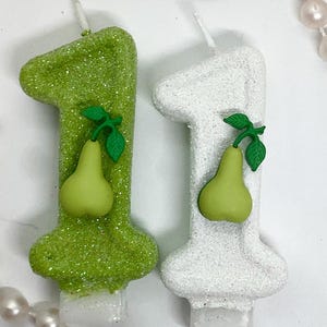 May include: Two number one shaped candles, one in lime green and one in white, both covered in a sparkling glitter texture. Each candle features a small, smooth, light green pear decoration with a green stem and two leaves attached to the front.