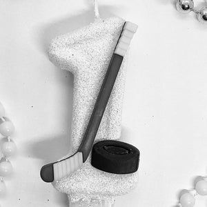 May include: A white number one candle with glitter, a hockey stick, and a black puck. The candle is surrounded by white and silver beaded garlands, creating a celebratory theme.