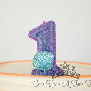 K&ouml;nnte beinhalten: Eine glitzernde lilafarbene Kerze in Form der Zahl eins mit einer blauen Muschel auf einem mehrschichtigen Kuchen in Wei&szlig; und Orange. Der Kuchen ist mit dem Text "Once Upon A Time Tutus" verziert.