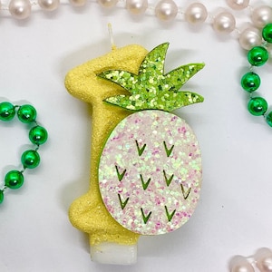 Op de afbeelding: Een gele ananasvormige kaars met groene bladeren en witte glitteraccenten. De kaars is in de vorm van het nummer één.