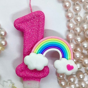 May include: Pink glitter candle shaped like the number one with a rainbow and clouds on top. The candle is surrounded by a string of pearls.
