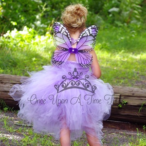 May include: A young girl wearing a lavender tulle tutu and butterfly wings. The wings are purple and white with a sparkly design. The girl is standing in a grassy area with a log behind her. The tutu has the text "Once Upon A Time Tutus" printed on it.