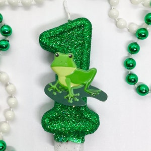 May include: Green glitter birthday candle shaped like the number one with a green frog on top. The candle is surrounded by green and white beads.