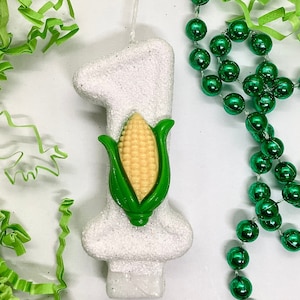 May include: White number one candle with a green and yellow corn cob design. The candle is decorated with glitter.