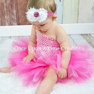 May include: A baby girl wearing a pink tulle tutu skirt and a white flower headband with a pink gem. The headband is on a pink elastic band. The baby is sitting on a white fluffy surface.