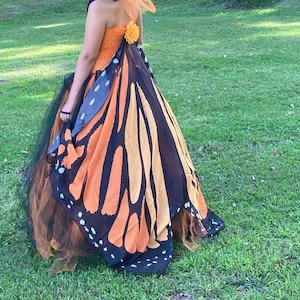 K&ouml;nnte beinhalten: Ein tr&auml;gerloses Kleid mit einem orangefarbenen, strukturierten Oberteil und einer kleinen orangefarbenen Blumenapplikation am R&uuml;cken. Der Rock besteht aus Lagen aus schwarzem T&uuml;ll und einem gro&szlig;en, flie&szlig;enden Umhang mit Schmetterlingsfl&uuml;gelmuster.