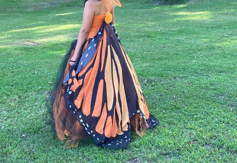 K&ouml;nnte beinhalten: Ein tr&auml;gerloses Kleid mit einem orangefarbenen, strukturierten Oberteil und einer kleinen orangefarbenen Blumenapplikation am R&uuml;cken. Der Rock besteht aus Lagen aus schwarzem T&uuml;ll und einem gro&szlig;en, flie&szlig;enden Umhang mit Schmetterlingsfl&uuml;gelmuster.