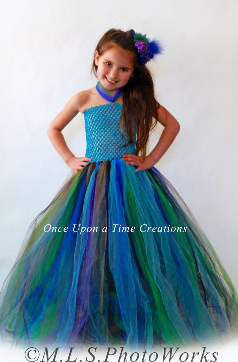 May include: A young girl wearing a turquoise and multi-colored tulle tutu dress with a crocheted bodice. The dress has a peacock feather design. The text "Once Upon a Time Creations" is visible on the dress.