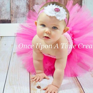 May include: A baby girl wearing a pink tulle tutu skirt and a white headband with a pink flower and a rhinestone. The baby is crawling on a wooden floor.