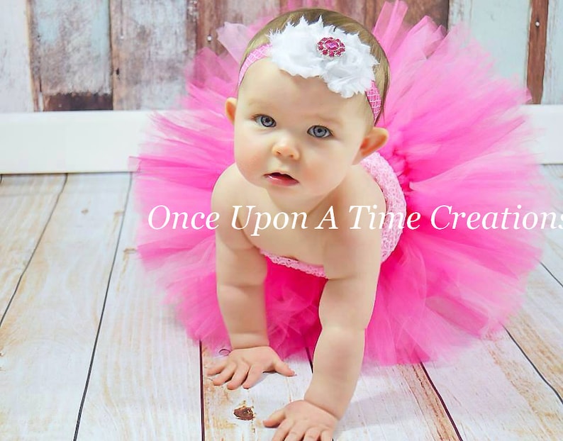 May include: A baby girl wearing a pink tulle tutu skirt and a white headband with a pink flower and a rhinestone. The baby is crawling on a wooden floor.