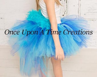 Puede incluir: Una camiseta blanca sin mangas combinada con una falda tutú de tul voluminosa de varias capas en tonos azul turquesa y azul real, con un detalle de plumas en la cintura. El texto Once Upon A Time Creations está superpuesto en la imagen.