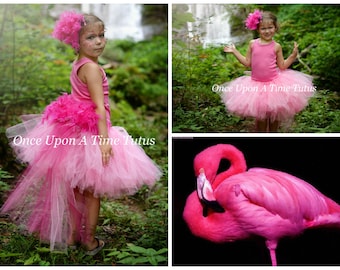 May include: A young girl wearing a pink tutu with a pink feather tail and a pink flower in her hair. The tutu is layered with tulle and has a pink feather trim. The girl is standing in a forest setting. The text "Once Upon A Time Tutus" is visible on the image.