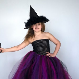 Puede incluir: Una ni&ntilde;a vestida de bruja para Halloween. Lleva un sombrero de bruja negro con plumas, un tut&uacute; de tul negro y morado y un corpi&ntilde;o de malla negra.