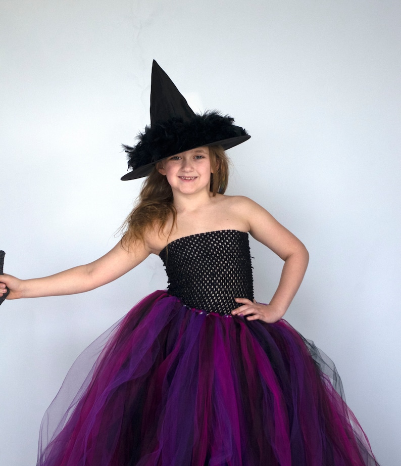 Puede incluir: Una ni&ntilde;a vestida de bruja para Halloween. Lleva un sombrero de bruja negro con plumas, un tut&uacute; de tul negro y morado y un corpi&ntilde;o de malla negra.