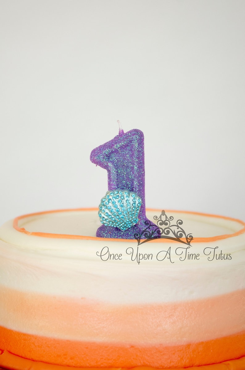K&ouml;nnte beinhalten: Eine glitzernde lilafarbene Kerze in Form der Zahl eins mit einer blauen Muschel auf einem mehrschichtigen Kuchen in Wei&szlig; und Orange. Der Kuchen ist mit dem Text "Once Upon A Time Tutus" verziert.