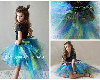 Op de afbeelding: Een pauw-thema tule tutu rok in tinten blauw, groen en paars met een lange sleep. De rok heeft een zwarte tailleband en is versierd met pauwenveren. De tekst "Once Upon A Time Tutus" is op de rok gedrukt.