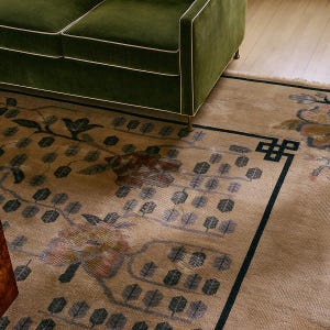 Puede incluir: Una alfombra beige con un diseño floral y de hojas en tonos verdes, marrones y azules. La alfombra tiene un borde verde oscuro y es parcialmente visible debajo de un sofá de terciopelo verde.