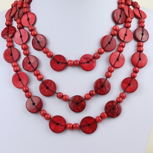 Puede incluir: Un collar de varias vueltas hecho de cuentas de madera roja. Las cuentas son redondas y tienen una superficie ligeramente texturizada. El collar está en capas y tiene un estilo grueso y bohemio.