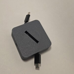May include: A square, dark gray charging hub with a textured, speckled finish. The device features a central diagonal slot and two black USB-C cables extending from opposite sides of the unit, designed for connecting and powering electronic devices.