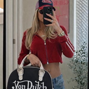 以下が含まれることがあります： 白のハンドルとシルバーのバックルが付いた、黒と白のVon Dutchハンドバッグ。赤いジップアップジャケット、白いストライプ、Von Dutchキャップ、サングラスをかけた人の前にバッグが持たれています。