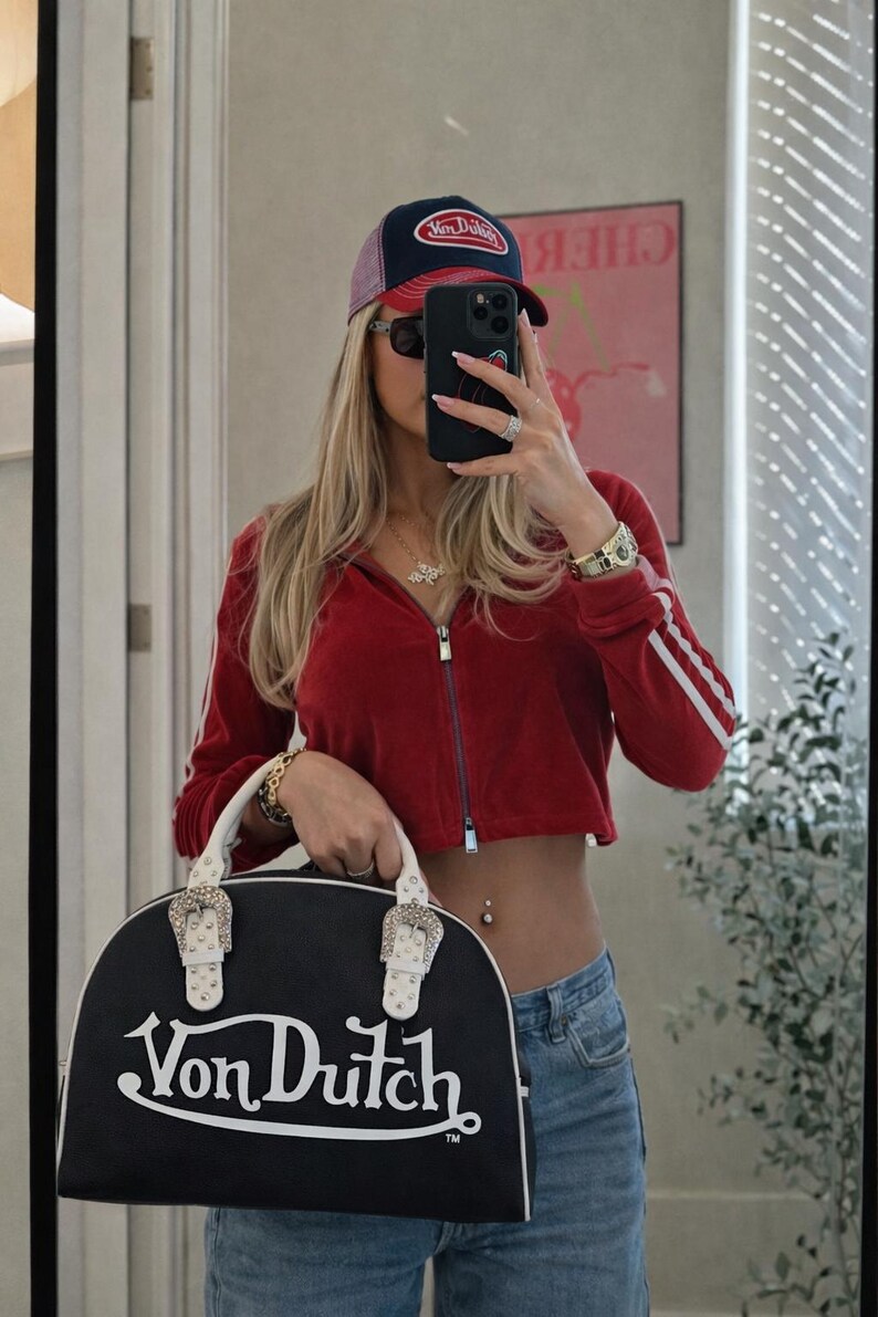 以下が含まれることがあります： 白のハンドルとシルバーのバックルが付いた、黒と白のVon Dutchハンドバッグ。赤いジップアップジャケット、白いストライプ、Von Dutchキャップ、サングラスをかけた人の前にバッグが持たれています。
