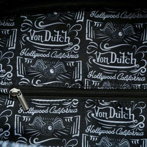 以下が含まれることがあります： 「Von Dutch Hollywood California」のテキストが入った、白と黒の模様の生地。 デザインには、翼のある目のグラフィックと装飾的な要素が含まれています。 銀色のジッパーが見えます。