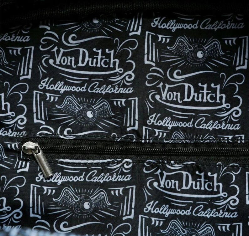 以下が含まれることがあります： 「Von Dutch Hollywood California」のテキストが入った、白と黒の模様の生地。 デザインには、翼のある目のグラフィックと装飾的な要素が含まれています。 銀色のジッパーが見えます。