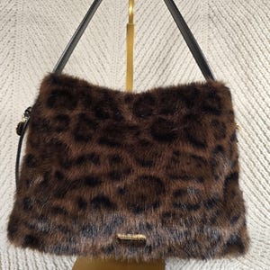 May include: A brown and black leopard print faux fur handbag featuring a structured rectangular body, a single black shoulder strap, and a small metallic logo plaque centred at the bottom edge of the front panel of the bag.