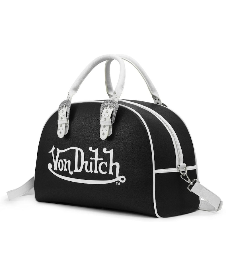 以下が含まれることがあります： 白文字で「Von Dutch」と書かれた黒と白のダッフルバッグ。バッグには白いハンドル、白いショルダーストラップ、シルバーカラーのバックルが付いています。バッグの高さは約25cm、幅は約46cmです。