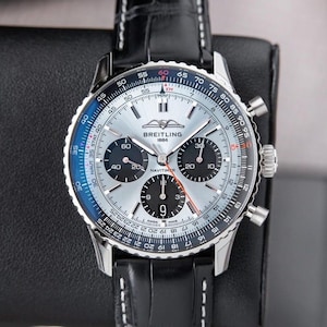 Könnte beinhalten: Eine Breitling Navitimer Uhr mit silbernem Zifferblatt, schwarzen Hilfszifferblättern und einem schwarzen Lederarmband. Die Uhr hat eine drehbare Lünette mit blauen und schwarzen Markierungen. Der Markenname ist sichtbar.